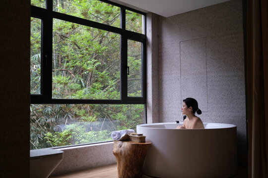 Woman relaxes in a bathtub next to a large window with views 