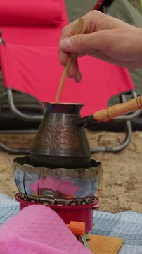 A person stirs fresh coffee inside an ornate copper cezve on a portable gas stove at a campsite. Concept of outdoor lifestyle, rustic morning routine, relaxation, and camping adventure.