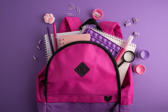 Bright Pink Backpack Filled With School Supplies And Stationery On A Purple Background