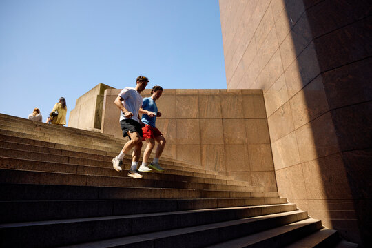 Two individuals run down the stairs in a sunny outdoor setting.