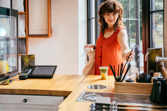 Cheerful middle-aged woman picking up colorful iced matcha drink at specialty cafe counter