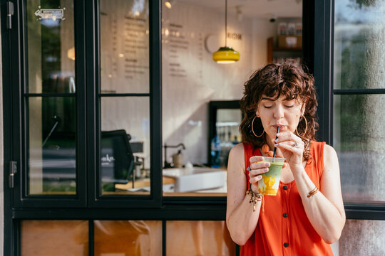 Relaxed middle-aged woman enjoying colorful iced matcha drink outside specialty cafe