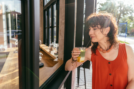 Relaxed middle-aged woman holding iced matcha by open cafe window on sunny summer day
