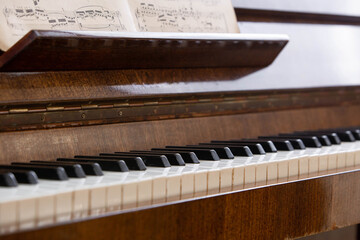 Close-up of a vintage upright piano with black and white keys and open sheet music resting on the music stand. Warm natural lighting highlights the wooden texture, creating a timeless atmosphere  © NataSao