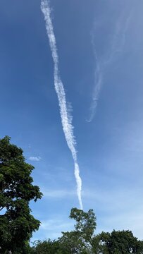Long aircraft contrail in blue sky above green trees