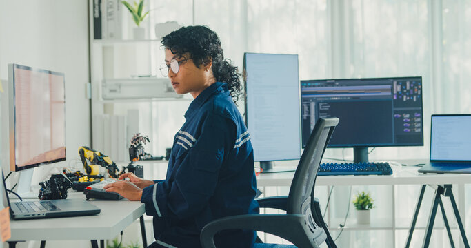 Young African American female robotics engineer coding AI software for robotic arm in laboratory. Embedded system, firmware testing, machine learning, industrial automation, future technology concept.