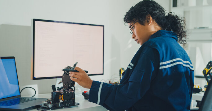 Young African American female robotics engineer coding AI software for robotic arm in laboratory. Embedded system, firmware testing, machine learning, industrial automation, future technology concept.