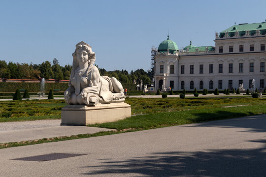 Statua monumentale di una sfinge in pietra nei giardini barocchi con la facciata del Palazzo del Belvedere Superiore sullo sfondo, famosa attrazione storica a Vienna, Austria.