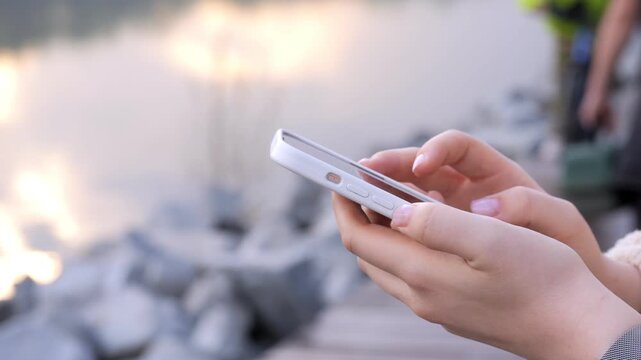 Close-up of hands using a smartphone near water. Blurred cyclist in background. Person focused on phone, calm and relaxed. Ideal for travel, tech, or leisure content