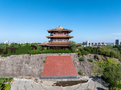 Traditional Chinese pavilion with golden roof stands atop rocky cliff with red staircase and plaza under clear blue sky