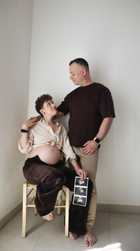 Happy pregnant woman sitting on chair holding ultrasound image with husband standing nearby