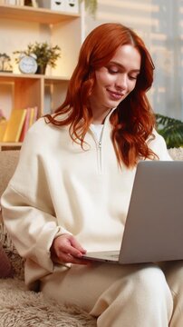 Young woman on home sofa opens laptop and starts typing with focused look as remote work begins. Adult lady on home sofa watches screen with calm attention and settles into familiar freelance rhythm.