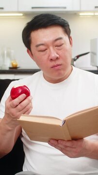 Asian man at home bites red apple enjoys juicy taste chews slow and smiles feeling fresh and awake. Chinese guy on sofa reads health tips in book marks page then eats more and feels fine and calm good