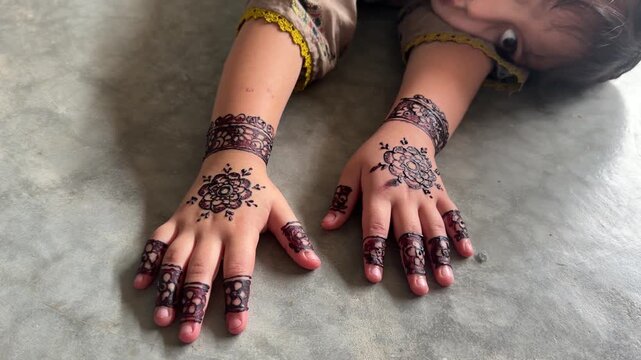 A young child proudly showing temporary henna designs on both hands and wrists, wearing patterned traditional dress against a black background.