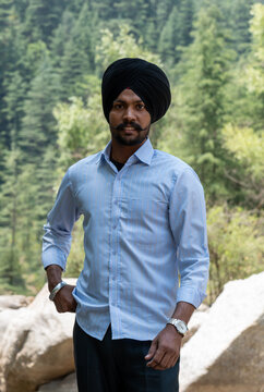A three-quarter portrait of an Indian Sikh man wearing a traditional black turban, a light blue pinstriped long-sleeve button-down shirt, and a wrist watch, looking at camera while standing outdoors.