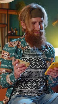 Bearded hippie man using credit bank card and smartphone while transferring money, purchases online shopping, order food delivery at home apartment indoors. Young redhead guy in evening room on couch