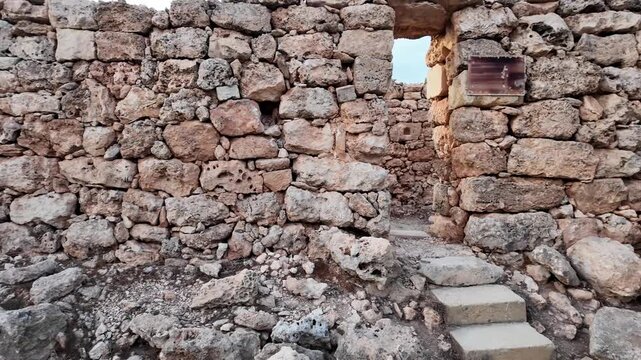 Ancient stone walls stand weathered and strong. Steps lead upward through a rustic doorway. Rough stones and gravel frame the scene. Sunlight glows softly behind the structure