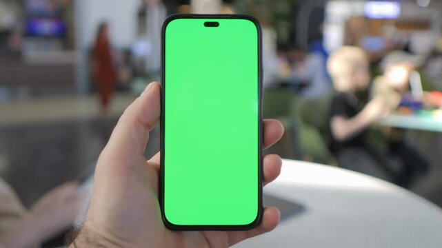 Hand holds phone with green screen in cafe. Blurred background shows people relaxing. Ideal for app demos or digital content. Neutral emotion, focused on device