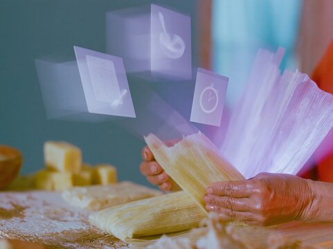 Close up of a womans hands folding a corn husk tamale on a weathered wooden table with glowing floating cooking icons for digital recipe and tradition concepts