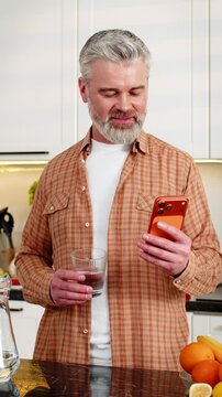 Middle-aged man smiles reading smartphone while drinking water in home kitchen during morning diet focus. Adult guy tracks hydration and healthy eating goals using mobile app indoors with calm mood