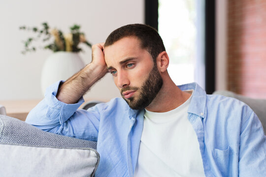 Young man feeling concerned and deep in thought while resting head on hand, sitting on a sofa in a modern living room