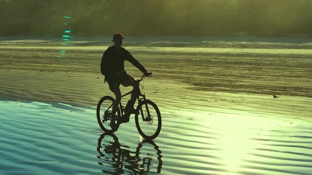 Silhouette of cyclist riding bicycle on reflective beach at golden sunrise. Peaceful coastal landscape with sunlight reflections. 4K video.
