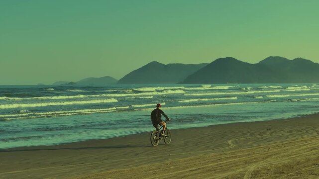 Cyclist riding along a tropical beach with mountains and ocean waves in the background. Scenic coastal landscape and active lifestyle. 4K video.