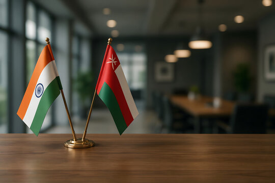 Crossed miniature flags of india and oman on a wooden table in a blurred office meeting room with warm lighting and dark chairs