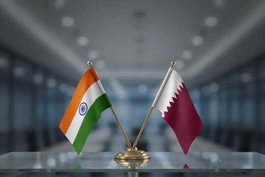 Miniature desk flags of india and qatar placed on a glass table in a blurred office setting, symbolizing international diplomatic relations