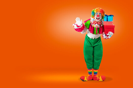 Funny clown with gifts friendly waving hand, man entertainer as Joker in a suit and wig, with clown whiteface makeup. Trickster, jester, pantomime, mime, professional actor at events, birthday party.