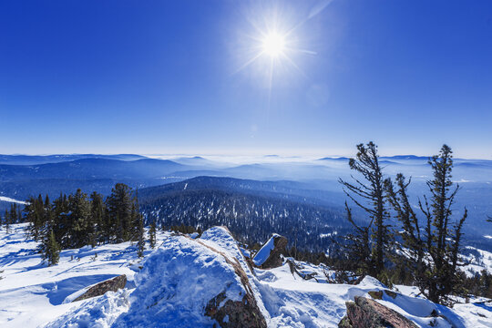 Panoramic view of mountain ridges in fog and sky in Sheregesh, Russia. Beauty white blue peaks Altay mountains fades in distance. Snow conifer forest and stones at bright winter sun on mount Ytuya.