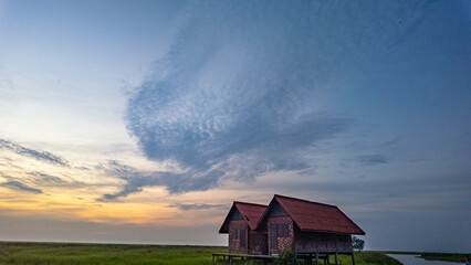 Twin traditional wooden houses stand peacefully on a green meadow beneath a dramatic sunset sky in Phatthalung, southern Thailand. A tranquil countryside landscape ideal for travel and nature themes. © Narong Niemhom