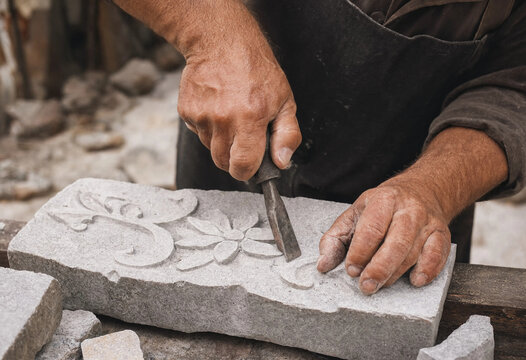 Craftsman meticulously carves an ornate floral pattern into a piece of stone using a sharp chisel and hammer.