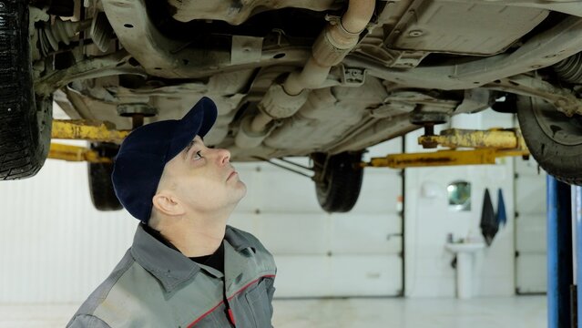Professional mechanic wearing protective cap checking underside of lifted vehicle in modern auto repair shop, focusing on chassis and exhaust system details