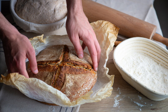 Freshly baked artisan sourdough bread with crispy crust held by baker hands. Rustic homemade loaf with banneton baskets, flour and baking tools on a wooden table.