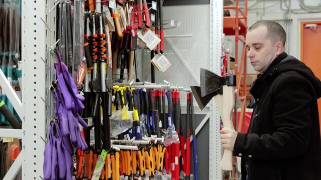 Male customer standing near display, comparing axe models with concentrated gaze, carefully checking specifications and weight for optimal woodcutting tool selection