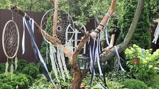 Dreamcatcher hanging on tree branch in garden. Decorative boho ornaments with ribbons for wedding ceremony. Outdoor mystical ethnic amulet in summer forest setting.