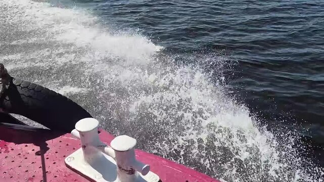 Motorboat moving fast through water creating white foam splashes. View from the red deck of a sailing vessel with a black fender over the side. Marine adventure.