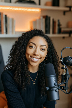 A lifestyle attractive woman creator sitting in a beautifully designed, minimalist home office, a podcast recording studio, by a microphone.