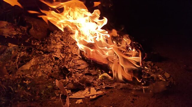 Bright orange flames rising from burning debris at night creates a warm glow with embers and smoke outdoors