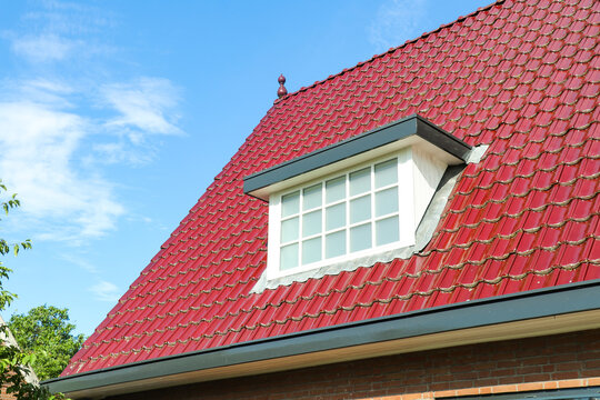 White dormer window on house with red clay roof tiles aginst blue sky. Residential architecture featuring pitched roof, rooftop roofing materials on suburban home.