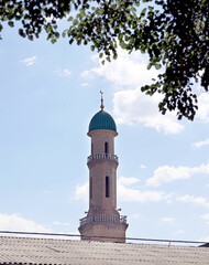Obraz premium Traditional mosque minaret rising above rooftops beneath a blue sky framed by tree branches. Peaceful architectural scene reflecting Islamic heritage in Uzbekistan.