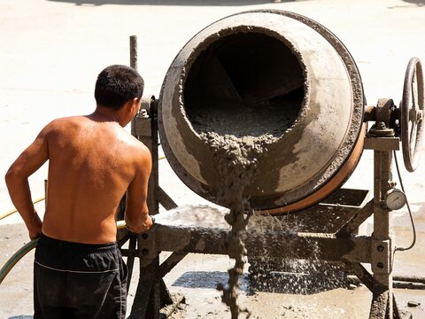 Shirtless construction worker operating a large rotating cement mixer while spraying water on grey concrete mixture