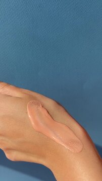Close up of female hands spreading applying liquid foundation on hand, make up routine. Creating a makeup swatch on blue background. Foundation application content, vertical lifestyle UGC content