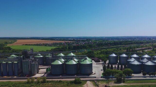 Dynamic aerial overview reveals rows of steel grain silos at modern agricultural storage facility near fields and railway tracks.