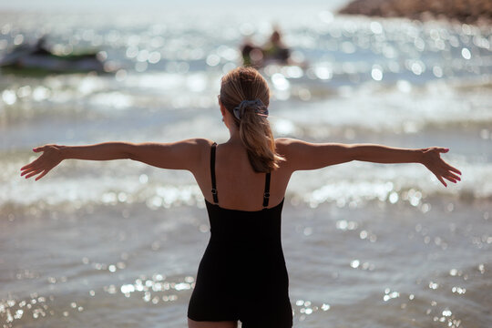 Back View Of Woman Opening Arms To Sea Enjoying Summer Vacation. Happy female tourist wearing black one-piece swimsuit stands on beach looking at ocean
