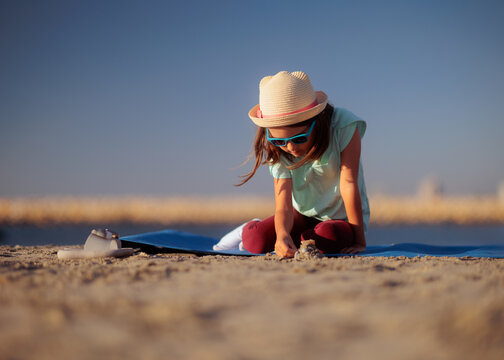 Cute Little Girl Wearing Hat And Sunglasses Playing On Beach. Happy young child enjoying summer vacation while sitting on mat playing with sand 