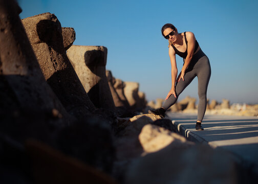 Fit Female Runner Stretching Legs On Wave-breaker Before Morning Jog. Active young woman wearing sunglasses leggings and top does pre-workout warm-up near ocean