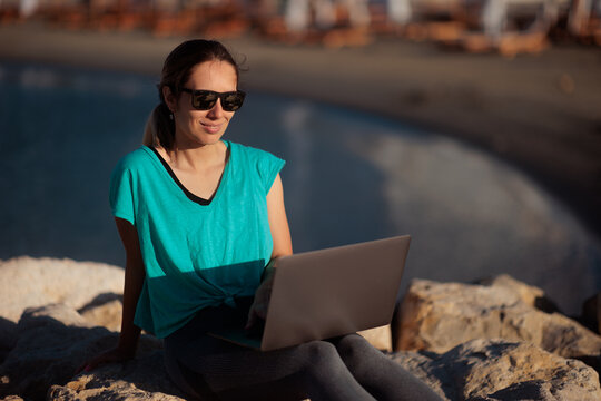 Happy Woman Wearing Sunglasses Working On Laptop Sitting On Wave breaker. Smiling freelancer enjoys remote business tasks during summer vacation near beautiful ocean