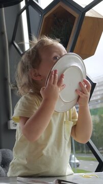 Hungry toddler with a stained t shirt sitting at a restaurant table, enthusiastically licking her empty plate clean after finishing a meal, showing great satisfaction and enjoyment